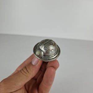 May include: A small, silver-colored ladybug-shaped object. The ladybug has a rounded body with small, circular indentations and a detailed head. The object is held in a person's hand against a plain white background.