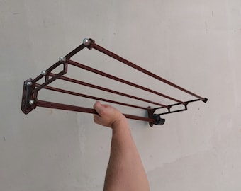 May include: A brown metal wall-mounted drying rack with five horizontal bars. The rack has decorative details and is mounted on a light-colored wall. The rack is being held by a person's hand.