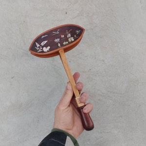 Puede incluir: Un instrumento musical de mano de madera con una parte superior redonda y decorada de color marrón y un mango de madera. La parte superior está decorada con un diseño floral y pájaros pequeños.