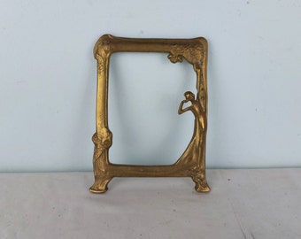 May include: A gold-toned metal picture frame with an Art Nouveau design featuring a woman's silhouette. The frame has a decorative floral motif and a raised edge.