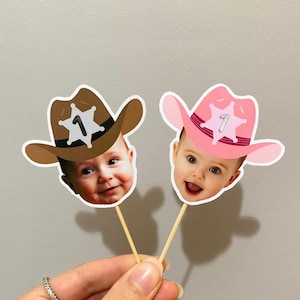 May include: Two cupcake toppers featuring children's faces with cowboy hats. The hats are brown and pink with a sheriff's star and the number "1" on them.