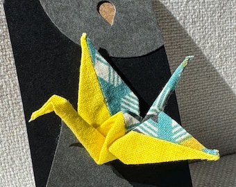May include: A yellow and teal origami crane displayed on a black card with a teardrop-shaped cutout. The crane is made of patterned fabric and is folded in the traditional Japanese style. The card has a gray top portion with a hole for hanging.