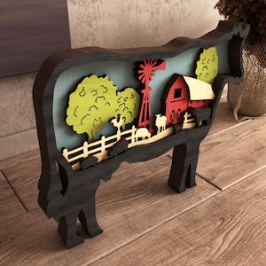 Op de afbeelding: Een houten decoratief stuk in de vorm van een koe met een boerderijscène. De scène omvat een rode schuur, een windmolen, bomen en boerderijdieren. Het stuk is in verschillende kleuren geschilderd en staat op een houten oppervlak.