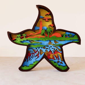 Pode incluir: Uma arte de parede de estrela do mar de madeira com um design em camadas. A estrela do mar é pintada em tons de marrom, verde, azul e laranja. O design apresenta uma cena de praia com palmeiras, um pôr do sol e um recife de coral.