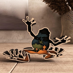 May include: A wooden frog-shaped art piece with a layered design. The frog is black with a detailed interior scene of a landscape with palm trees and an orange and green color scheme. The art piece is on a wooden surface.