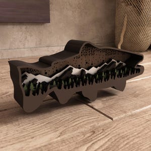 May include: A brown wooden sculpture in the shape of a fish, featuring a layered design with a mountain and forest scene. The sculpture has a detailed, cut-out pattern on the top and is displayed on a wooden surface.