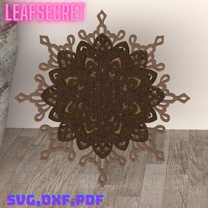 May include: A brown and green layered mandala design cut from wood. The design is in the shape of a snowflake with intricate details. The text "LEAFSECRET" is at the top of the image and "SVG, DXF, PDF" is at the bottom.