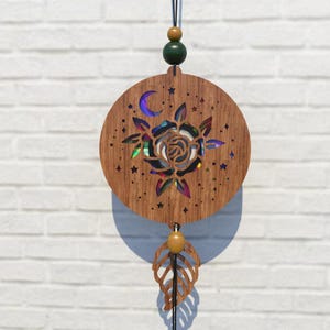 May include: Wooden wall hanging featuring a laser-cut rose design with a crescent moon and stars. The rose has iridescent accents. A leaf-shaped charm hangs below. The wall decor is suspended by a black cord with wooden beads.