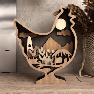 May include: Wooden hen-shaped decorative piece with a layered design. The interior features a landscape with mountains, a rooster, and a sun. The piece is made of light brown wood and stands on a flat surface.