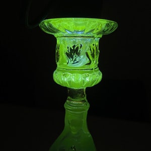 Op de afbeelding: Een groene glazen kandelaar met een decoratief ontwerp. De kandelaar is gemaakt van uraniumglas en gloeit onder een zwarte lamp.