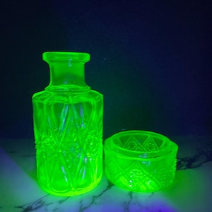 Può includere: Una bottiglia in vetro verde neon fluorescente e una piccola ciotola in vetro abbinata, entrambe caratterizzate da un motivo testurizzato a rombi e fiori. Il vetro emette una luce brillante sotto i raggi ultravioletti, evidenziando i dettagli geometrici in rilievo.