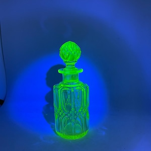 Peut inclure: Flacon de parfum en verre vert néon avec un bouchon décoratif. La bouteille a une forme rectangulaire avec des motifs gravés. Le bouchon est rond avec un design texturé. La bouteille est éclairée par une lumière bleue.