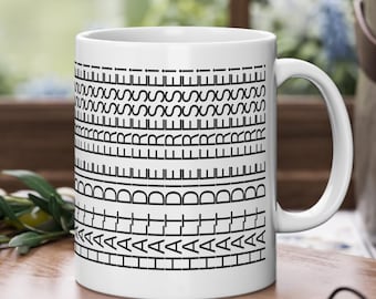 Puede incluir: Taza de café de cerámica blanca con un patrón geométrico negro. El diseño presenta varias bandas horizontales de líneas, curvas y formas. La taza está sobre una tabla de cortar de madera con aceitunas y utensilios en el fondo.