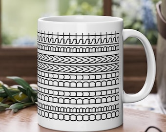 Puede incluir: Taza de cerámica blanca con un patrón geométrico negro. El diseño presenta bandas horizontales de varias formas, incluyendo líneas, chevrones y semicírculos. La taza tiene un asa curva.