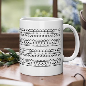 Könnte beinhalten: Weiße Keramiktasse mit einem schwarzen geometrischen Muster. Das Design zeigt sich wiederholende Reihen verschiedener Formen, darunter Bögen, Dreiecke und Kreise. Die Tasse hat einen gebogenen Griff.