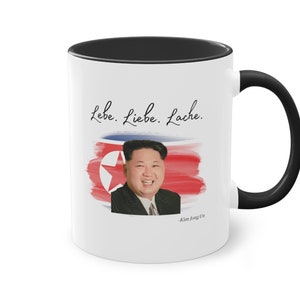 Op de afbeelding: Witte keramische koffiemok met een zwarte rand en handvat. De mok heeft een aquarelimage van de Noord-Koreaanse vlag met een portret van Kim Jong Un. De tekst "Lebe. Liebe. Lache." staat boven de afbeelding, en de tekst "Kim Jong Un" staat onder de afbeelding.