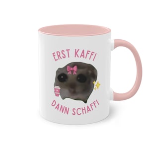 Könnte beinhalten: Weiße Keramik-Tasse mit rosa Innenraum und Henkel. Die Tasse zeigt ein Cartoon-Bild einer Kreatur mit rosa Schleife und Kaffeetasse sowie den Text "ERST KAFFI DANN SCHAFFI" in Rosa.