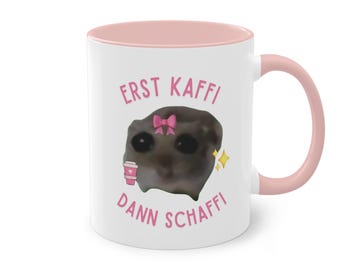May include: White ceramic mug with a pink interior and handle. The mug features a cartoon image of a creature with a pink bow and a coffee cup, along with the text "ERST KAFFI DANN SCHAFFI" in pink.