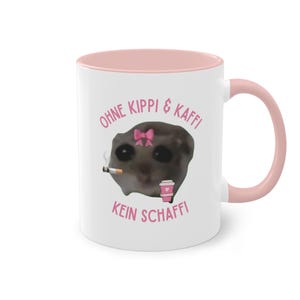 Könnte beinhalten: Eine weiße Keramiktasse mit hellrosa Innenseite und Henkel. Auf der Vorderseite ist ein Hamster mit rosa Schleife abgebildet, der eine Zigarette und einen rosa Kaffeebecher hält, mit dem rosa Text OHNE KIPPI & KAFFI KEIN SCHAFFI.