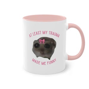 Könnte beinhalten: Weiße Keramiktasse mit rosafarbenem Rand und Henkel. Die Tasse zeigt ein Cartoon-Bild eines Lemurs mit einer rosafarbenen Schleife und dem Text "Zumindest mein Trauma hat mich lustig gemacht".