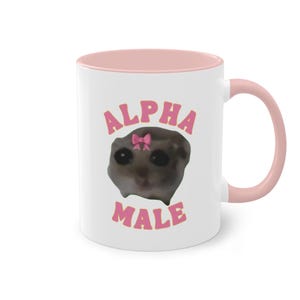 Könnte beinhalten: Weiße Keramiktasse mit rosafarbenem Rand und Henkel. Die Tasse zeigt ein Cartoon-Bild einer Katze mit einer rosa Schleife auf dem Kopf. Der Text "ALPHA MALE" ist in rosa Buchstaben auf die Tasse gedruckt.