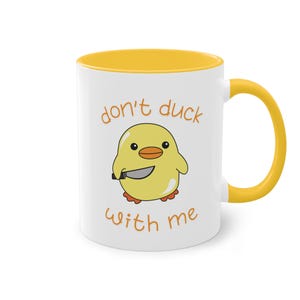 Könnte beinhalten: Weiße Keramiktasse mit gelbem Rand und Henkel. Die Tasse zeigt ein gelbes Cartoon-Küken, das ein Messer hält, und den Text "don't duck with me" in Orange.