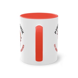 May include: White ceramic coffee mug with a red rim and handle. The mug has a black and red graphic on one side that says "Pyromanik ist doch kein Verbrechen" and a black and red graphic on the other side that says "Pyromanik".