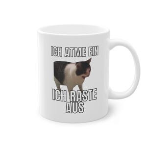 Op de afbeelding: Witte keramische mok met een zwart-witte kat afbeelding en de tekst "ICH ATME EIN ICH RASTE AUS" erop gedrukt.