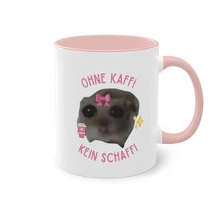 Pode incluir: Caneca de cerâmica branca com interior e alça rosa. A caneca apresenta uma imagem de desenho animado de uma criatura com um laço rosa e o texto alemão "OHNE KAFFI KEIN SCHAFFI" em rosa, junto com gráficos de xícara de café e estrelas.