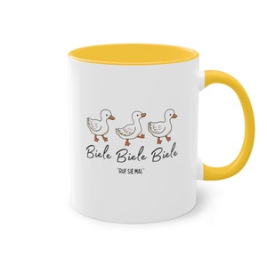Könnte beinhalten: Weißer Keramikbecher mit gelbem Innenraum und Henkel. Der Becher zeigt ein skurriles Design von drei Cartoon-Enten, die in einer Reihe gehen, mit dem Text "Biele Biele Biele" und "RUF SIE MAL" darunter.