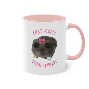Könnte beinhalten: Weiße Keramiktasse mit rosafarbenem Rand und Henkel. Die Tasse zeigt ein Cartoon-Bild eines kleinen Tieres mit einer rosa Schleife, das einen Bleistift und ein Buch hält. Der Text "ERST KAFFI DANN THERAPI" ist in rosa auf die Tasse gedruckt.