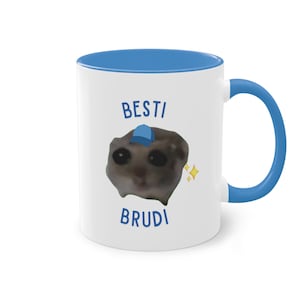 Könnte beinhalten: Weiße Keramiktasse mit blauem Rand und Henkel. Die Tasse zeigt ein Cartoon-Bild eines kleinen Tieres, das eine blaue Baseballkappe trägt. Der Text "BESTI BRUDI" ist in Blau auf die Tasse gedruckt.