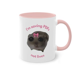 Könnte beinhalten: Eine weiße Keramiktasse mit rosafarbenem Rand und Henkel. Die Tasse zeigt ein Cartoon-Bild eines traurigen Tieres mit einer rosafarbenen Schleife auf dem Kopf. Der Text "I'm saving PDFs not lives" ist in Rosa auf die Tasse gedruckt.