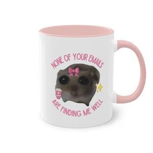 Op de afbeelding: Een witte keramische mok met een roze rand en handvat. De mok heeft een cartoonafbeelding van een kat met een roze strik en een koffiebeker. De tekst op de mok luidt "None of your emails are finding me well."