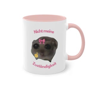 Könnte beinhalten: Weiße Keramiktasse mit rosafarbenem Rand und Henkel. Die Tasse zeigt ein Cartoon-Bild eines Lemurs mit einer rosafarbenen Schleife und den Text "Nicht meine Zuständigkeit" in rosa.