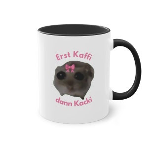 Könnte beinhalten: Weiße Keramik-Kaffeetasse mit schwarzem Rand und Henkel. Die Tasse zeigt ein Cartoon-Bild eines Lemurs mit einer rosa Schleife und dem Text "Erst Kaffee dann Kacki" in rosa.