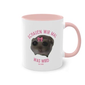 Könnte beinhalten: Weiße Keramiktasse mit rosafarbenem Rand und Henkel. Die Tasse zeigt ein Cartoon-Bild eines kleinen Tieres mit einer rosafarbenen Schleife auf dem Kopf. Der Text "Schauen wir mal was wird" ist in Rosa auf die Tasse gedruckt.