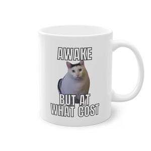 Könnte beinhalten: Weiße Keramiktasse mit einer Katze und dem Text "AWAKE BUT AT WHAT COST". Die Katze ist weiß mit schwarzen Markierungen. Der Text ist in fetter, schwarzer Schrift. Die Tasse hat einen gebogenen Griff.