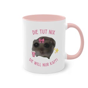 Könnte beinhalten: Weiße Keramiktasse mit rosafarbenem Rand und Henkel. Die Tasse zeigt ein Cartoon-Bild einer Katze mit einer rosa Schleife und einem rosa Kaffeetassen. Der Text "DIE TUT NIX DIE WILL NUR KAFFI" ist in rosa auf die Tasse gedruckt.
