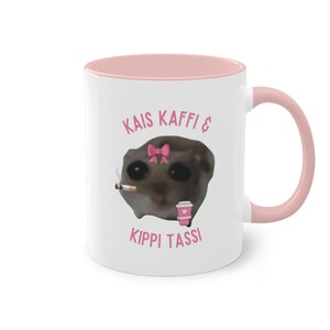 Könnte beinhalten: Weiße Keramik-Tasse mit rosa Innenraum und Henkel. Die Tasse zeigt ein Cartoon-Bild einer Kreatur mit rosa Schleife, Zigarette und Kaffeetasse. Der Text auf der Tasse lautet "KAIS KAFFI & KIPPI TASSI."