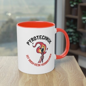 May include: White ceramic mug with a red rim and handle. The mug features a black and red illustration of a lit torch with the text "Pyrotechnik ist doch kein Verbrechen" around it.
