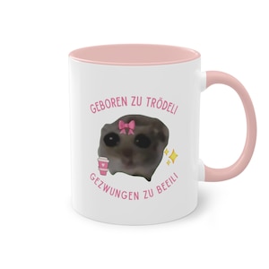 Könnte beinhalten: Weiße Keramiktasse mit rosafarbenem Rand und Henkel. Die Tasse zeigt ein Cartoon-Bild eines kleinen Tieres mit einer rosa Schleife und einer Kaffeetasse. Der Text auf der Tasse lautet "Geboren zu trödeli Gezwungen zu beeili".