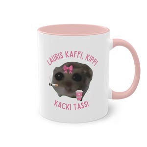 Könnte beinhalten: Eine weiße Keramiktasse mit rosafarbenem Rand und Henkel. Die Tasse zeigt ein Cartoon-Bild einer Katze mit einer rosa Schleife, die eine Zigarette und eine rosa Tasse Kaffee hält. Der Text "LAURIS KAFFI, KIPPI KACKI TASSI" ist in rosa auf die Tasse gedruckt.