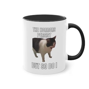 Könnte beinhalten: Weiße Keramik-Kaffeetasse mit schwarzem Rand und Henkel. Die Tasse zeigt ein Schwarzweißbild einer Katze mit dem Text "The Horrors Persist But So Do I".