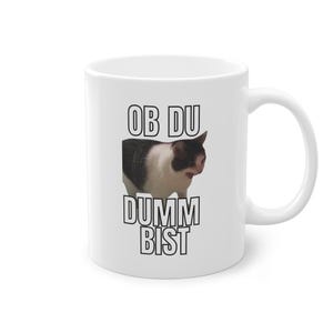 Könnte beinhalten: Weiße Keramik-Kaffeetasse mit einem Schwarzweiß-Katzenbild und dem aufgedruckten Text "OB DU DUMMY BIST".