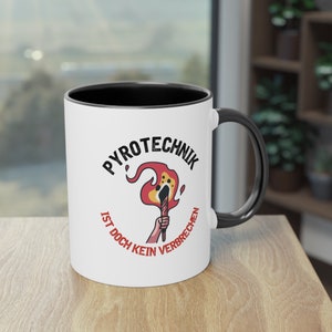 May include: White ceramic mug with a black rim and handle. The mug features a red and yellow illustration of a lit torch with the text "Pyrotechnik ist doch kein Verbrechen" in black.