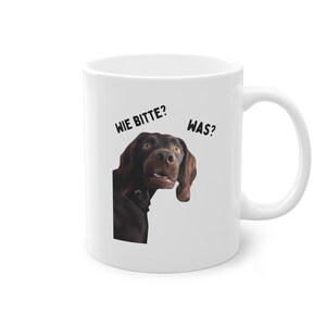 Könnte beinhalten: Weißer Keramikbecher mit einem überraschten braunen Hund. Die deutschen Sätze „WIE BITTE?“ und „WAS?“ sind in schwarzer Schrift aufgedruckt. Der Becher hat einen gebogenen Griff.