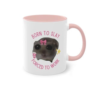Könnte beinhalten: Weiße Keramiktasse mit rosafarbenem Rand und Henkel. Die Tasse zeigt ein Cartoon-Bild einer Katze mit einer rosa Schleife und dem Text "Born to Slay Forced to Work".