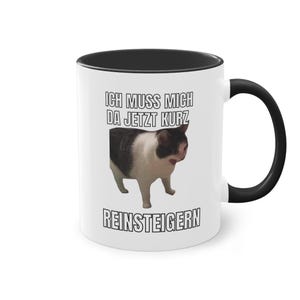 May include: White ceramic coffee mug with a black rim and handle. The mug features a black and white image of a cat with the text "Ich muss mich da jetzt kurz reinsteigen".