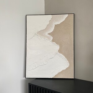May include: Abstract art painting in a black frame with white and beige textured paint. The painting is hanging on a wall above a black cabinet.